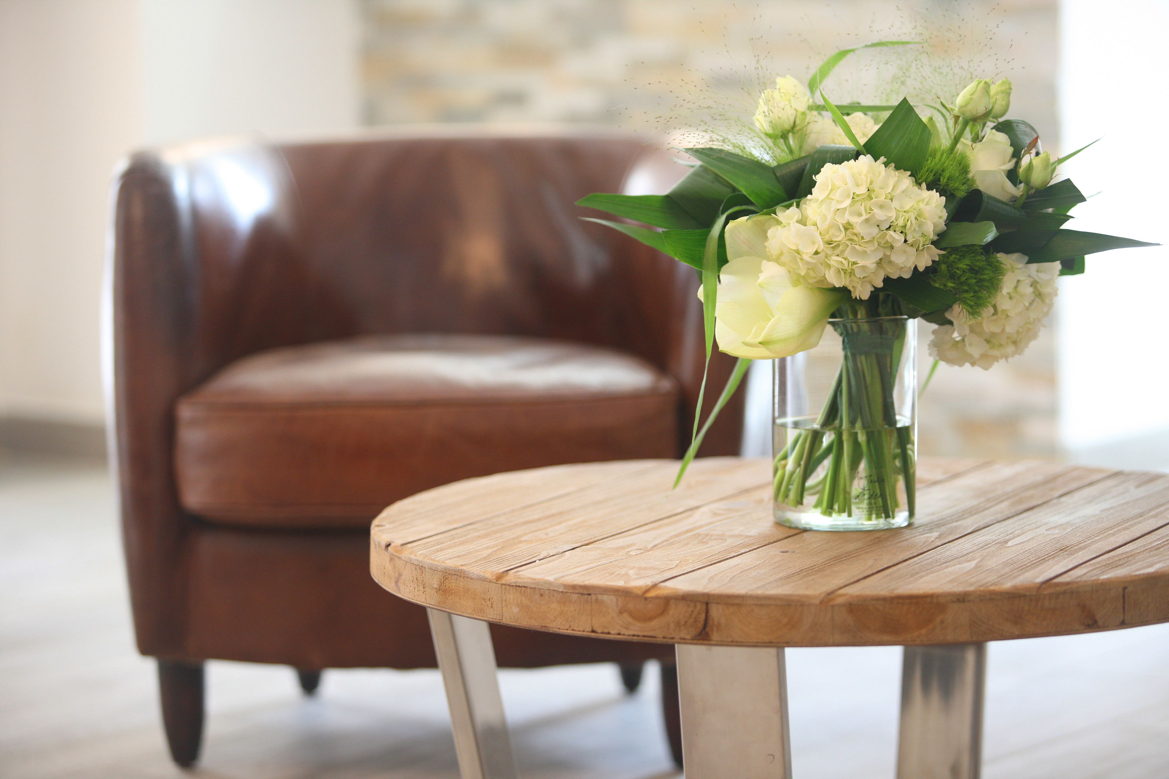 A wooden table with a clear glass vase containing white flowers and green foliage.
