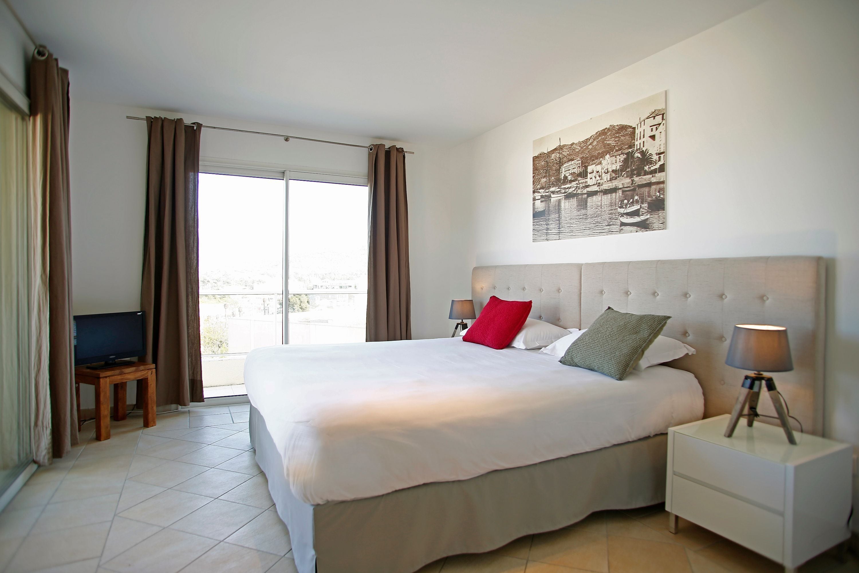 A hotel room with a large bed, a television on a small table, a nightstand with a lamp, and a balcony with curtains.