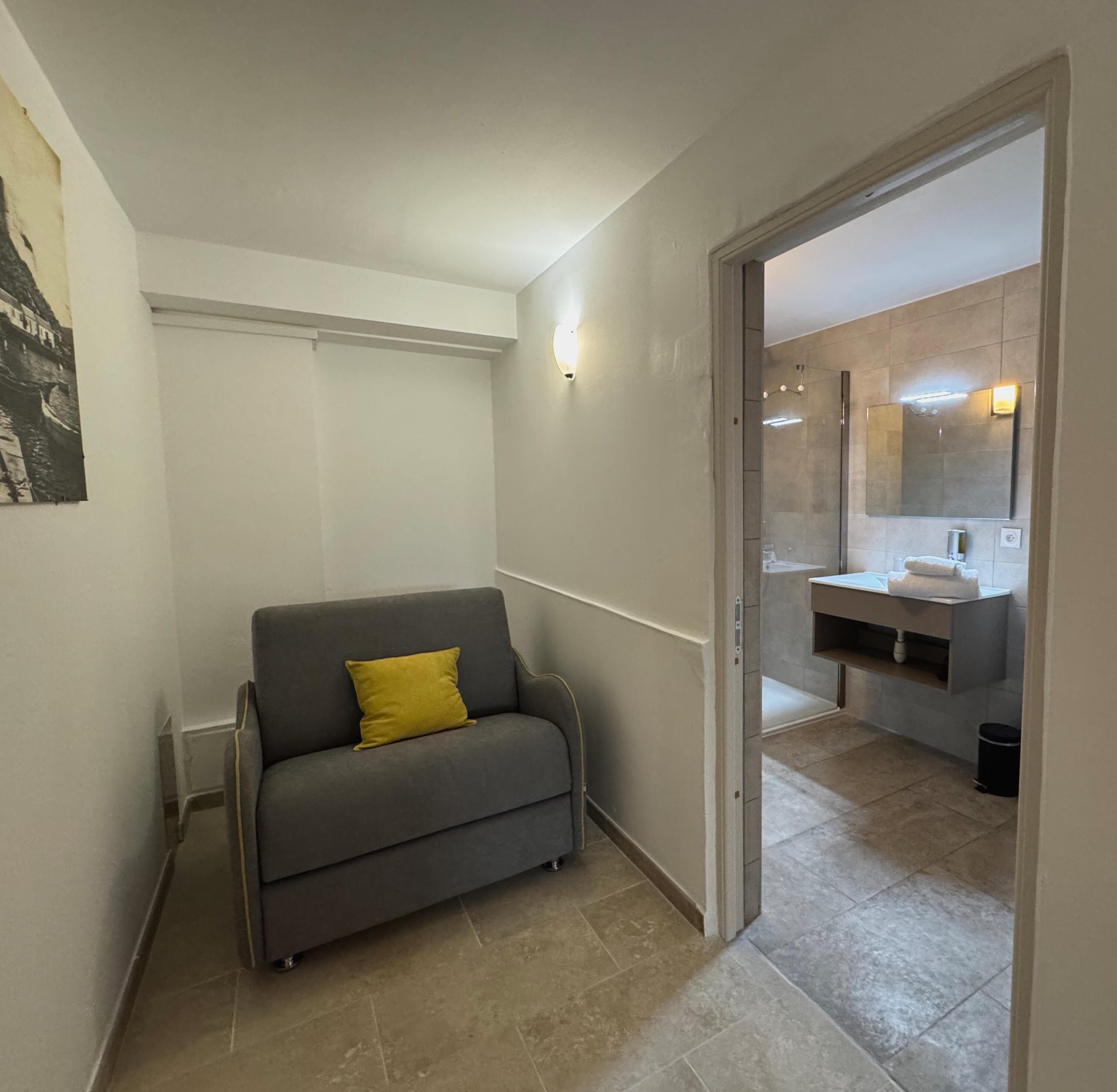 A modern, compact living space with a grey sofa and a yellow pillow, leading to a bathroom with a large mirror and a sink.