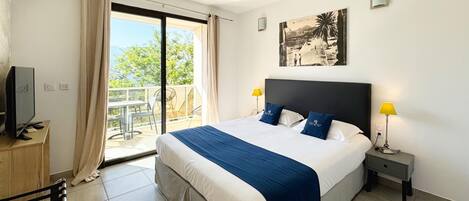 A hotel room with a large bed, a TV, a desk, and a balcony with a view of trees and a building.