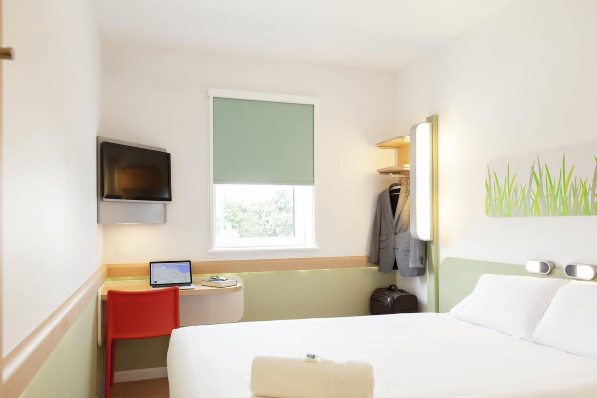 A hotel room with a bed, a desk with a laptop, a TV mounted on the wall, and a window with a view of trees.