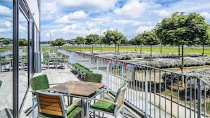 An outdoor seating area with tables and chairs, a metal railing, and a view of a landscaped garden with trees and bushes. - ibis budget Edinburgh Park (Edinburgh)
