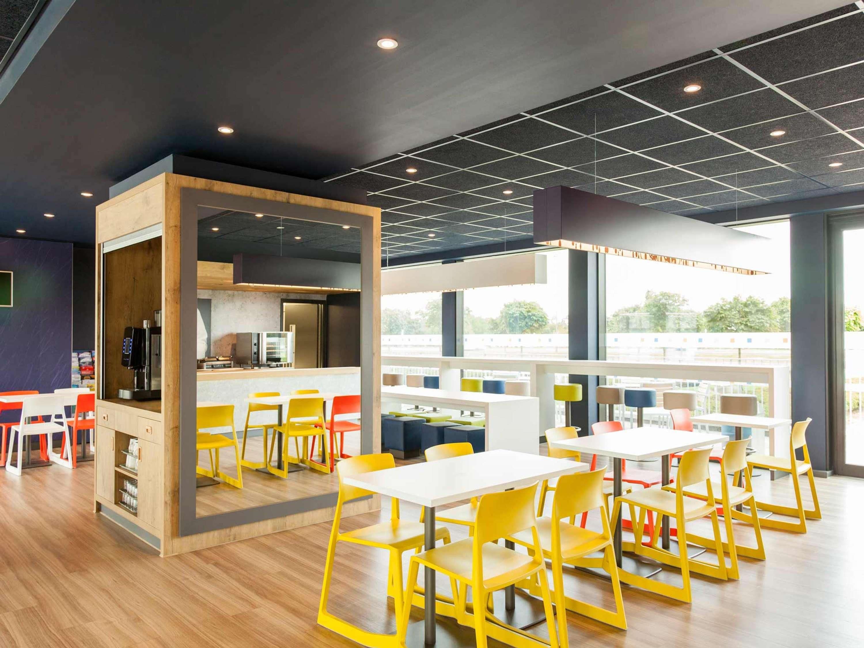 A modern dining area with yellow chairs, white tables, and a kitchen area with a coffee machine.