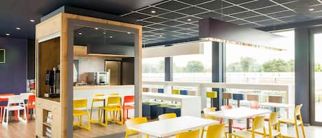 A modern dining area with yellow chairs, white tables, and a kitchen area with a coffee machine.