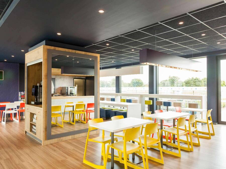 Een moderne eethoek met gele stoelen, witte tafels en een keukenhoek met een koffieapparaat.