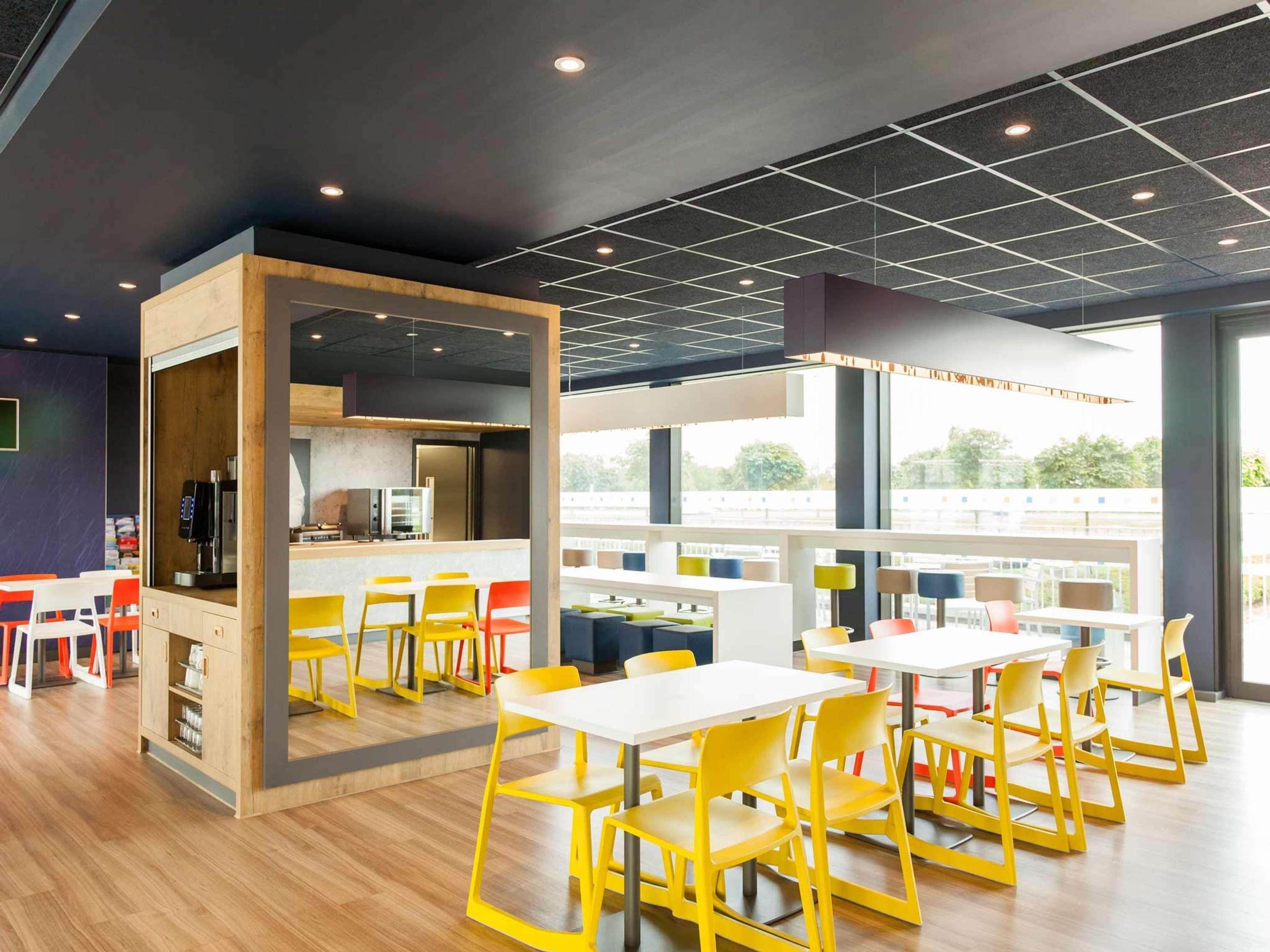 A modern dining area with yellow chairs, white tables, and a kitchen area with a coffee machine.