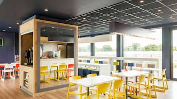 A modern dining area with yellow chairs, white tables, and a kitchen area with a coffee machine.
