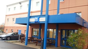 A building with a blue awning, a sign reading "Bourges", and a parking area with cars. - ibis budget Bourges (Le Subdray)
