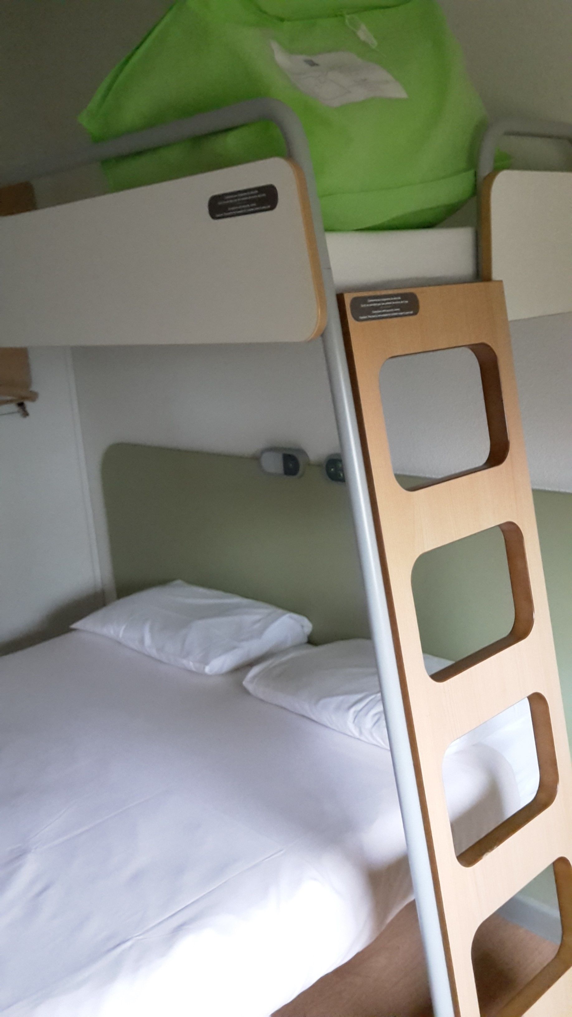 A bunk bed with a wooden ladder, white bedding, and a green bag on the top bunk.