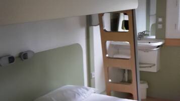 Ein Stockbett mit einem grünen Beutel auf der oberen Ebene, ein Nachttisch, ein Spiegel und ein kleines Regal mit einem Waschbecken.