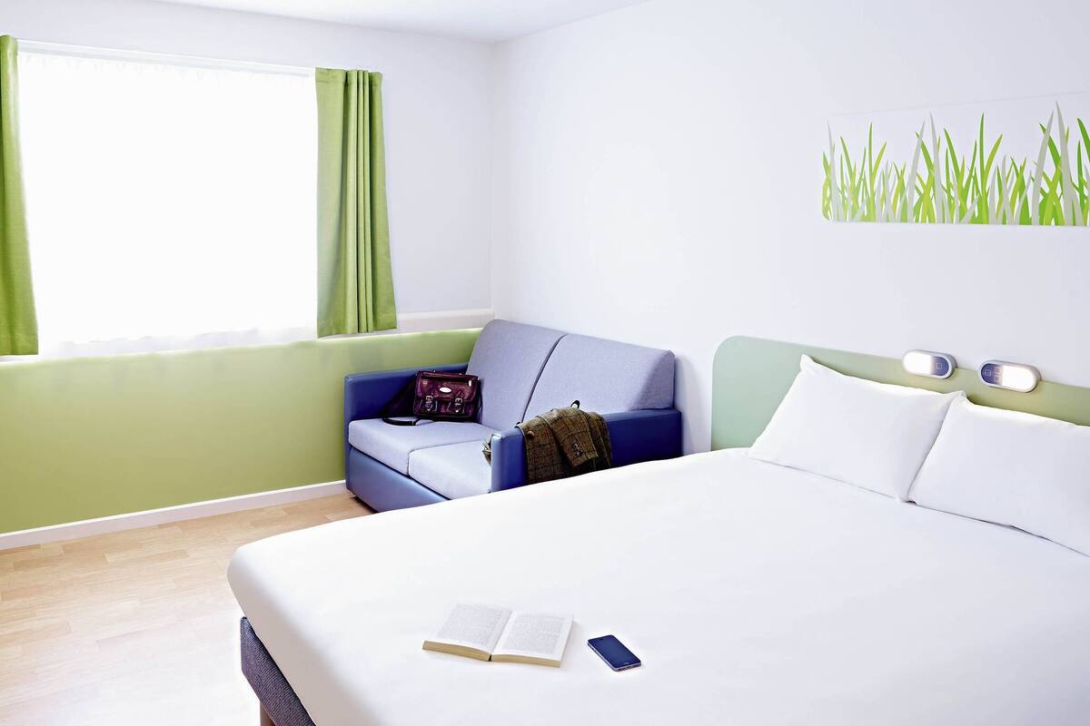 Une chambre d’hôtel avec un lit, un fauteuil bleu, un mur vert et une fenêtre avec des rideaux.