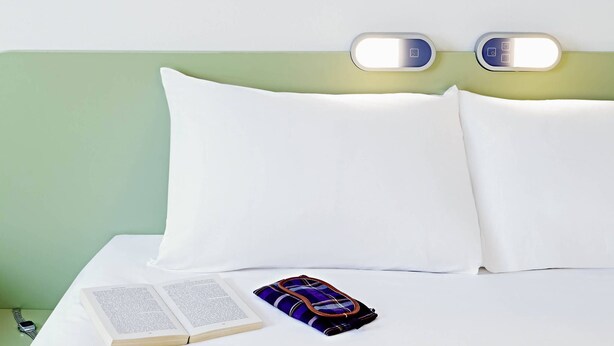 Un lit bien fait, avec des oreillers blancs et une tête de lit verte. On y voit un livre et un sac coloré.