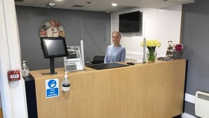 Un comptoir d’accueil avec une horloge, un panneau indiquant un tarif de stationnement réduit de moitié, du gel hydroalcoolique et un détecteur d’incendie.