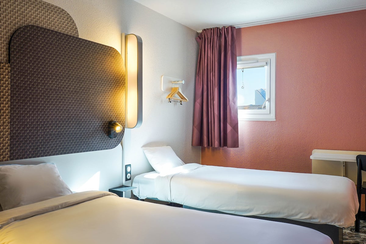 Une chambre d’hôtel avec deux lits, une lampe fixée au mur, une fenêtre avec des rideaux et une petite table avec une chaise.