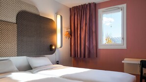 Une chambre d’hôtel avec un lit, une tête de lit, une fenêtre avec des rideaux et un luminaire fixé au mur.