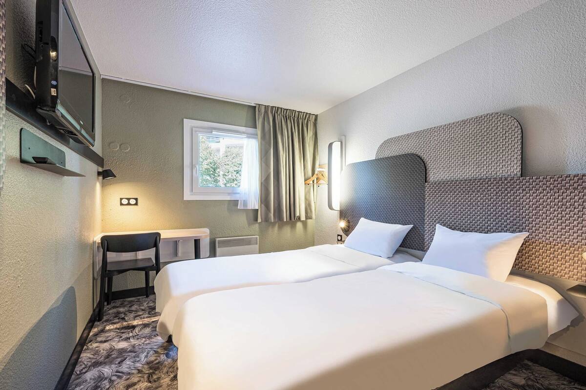 Habitació d'hotel amb dos llits, una televisió, una cadira, una finestra amb cortines i una làmpada penjada a la paret.