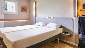 A hotel room with two beds, a small bedside table, a wall-mounted shelf, and a window.