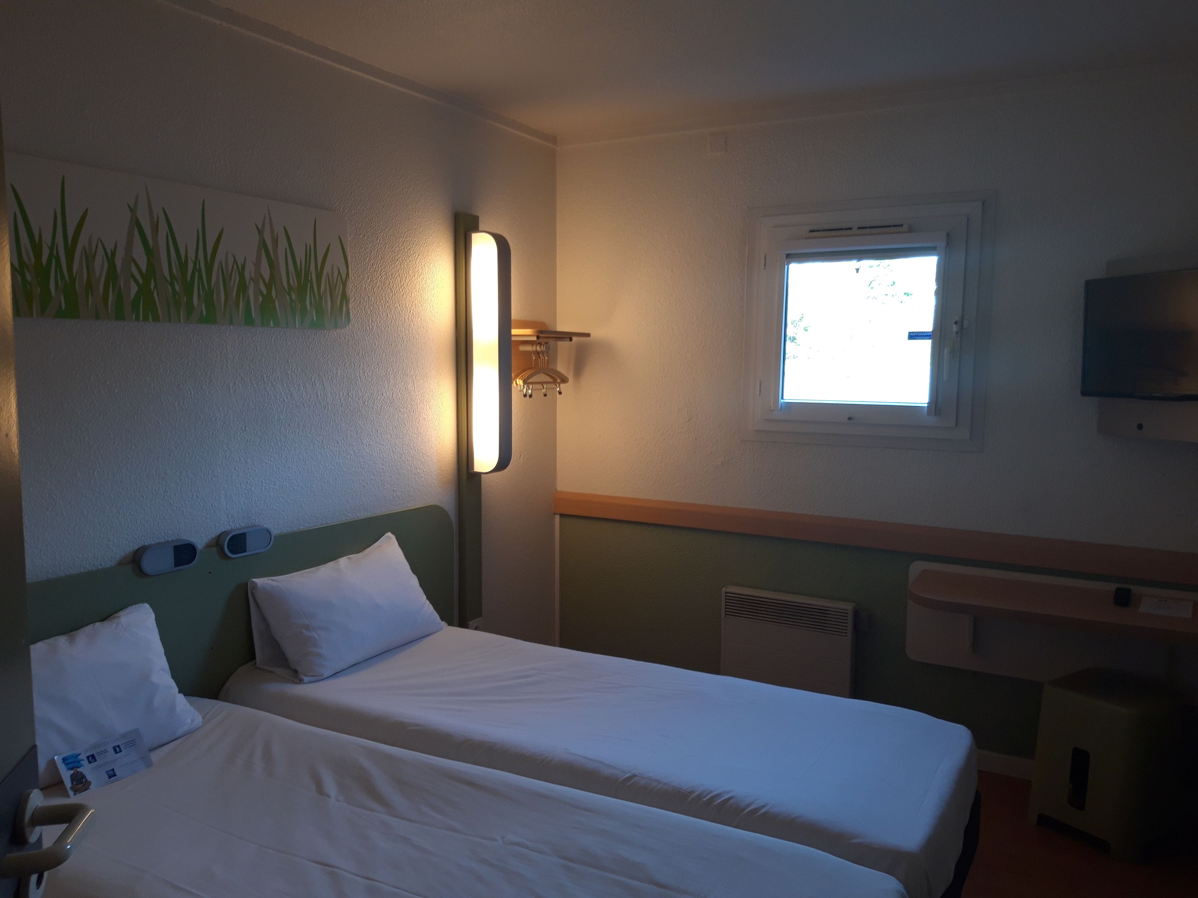 A hotel room with two beds, a TV mounted on the wall, a window with a view, and a wall-mounted light fixture.