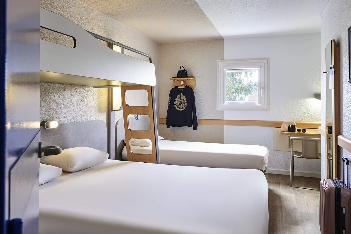 Habitación de hotel compacta con dos camas individuales, un escritorio pequeño, una ventana con vistas y una cama individual superior.