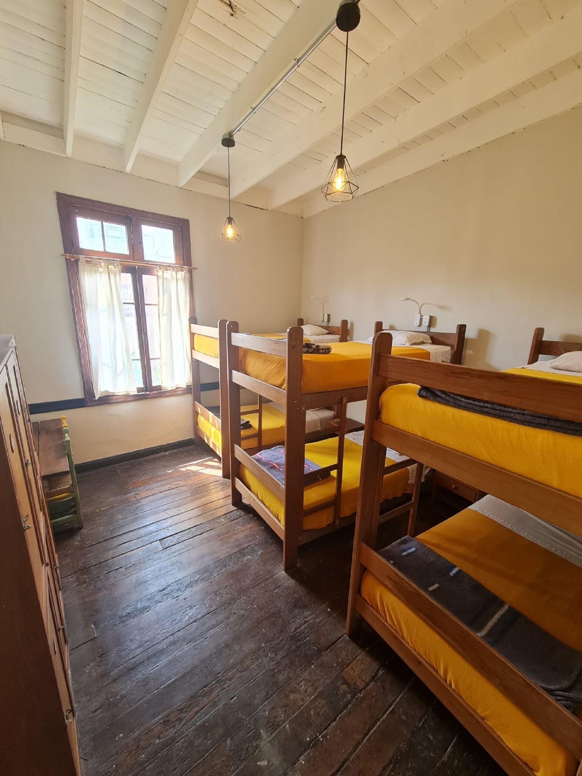 A dormitory room with bunk beds, wooden floors, a window with curtains, and hanging pendant lights.