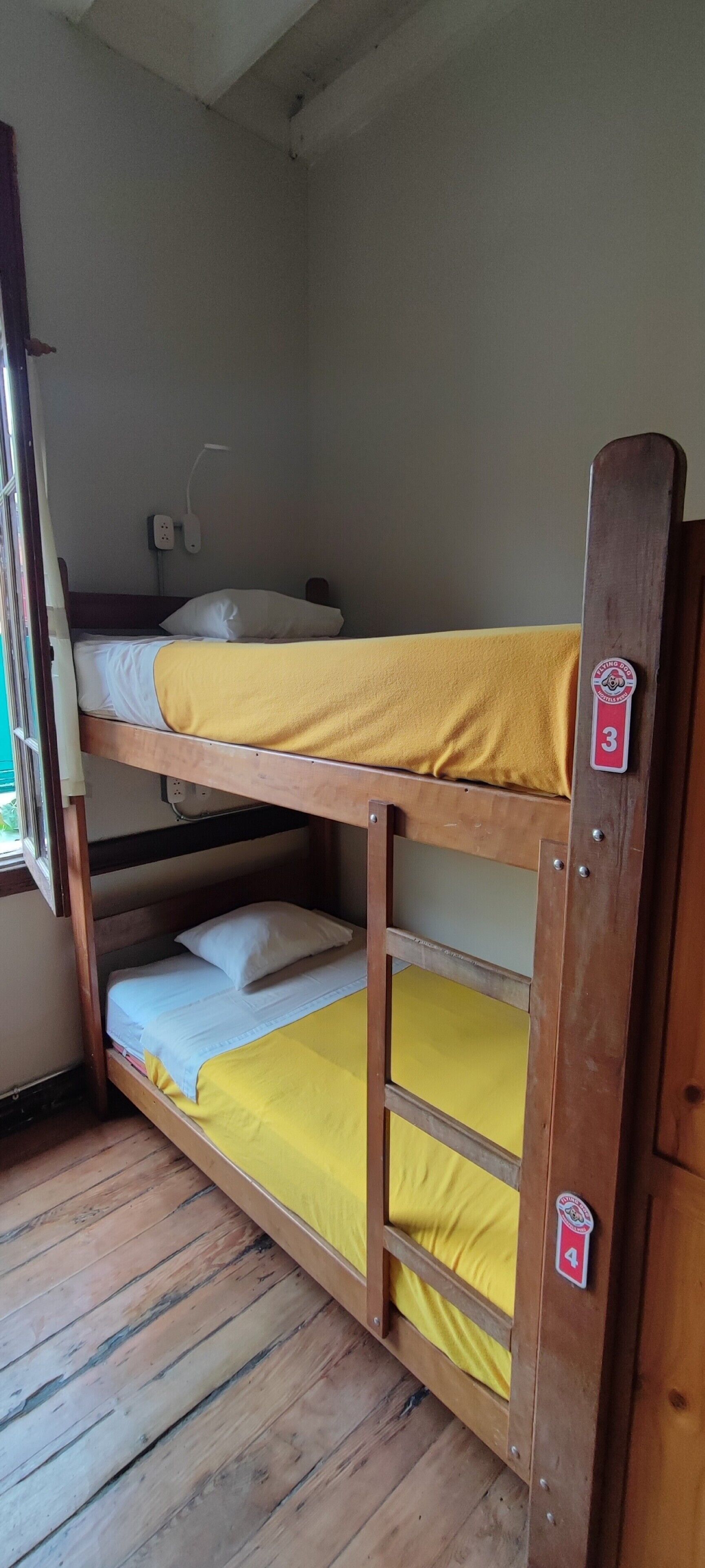 A bunk bed with three levels, each with a yellow mattress and white pillows. There is a window on the left side, and a wall socket above the top bunk.
