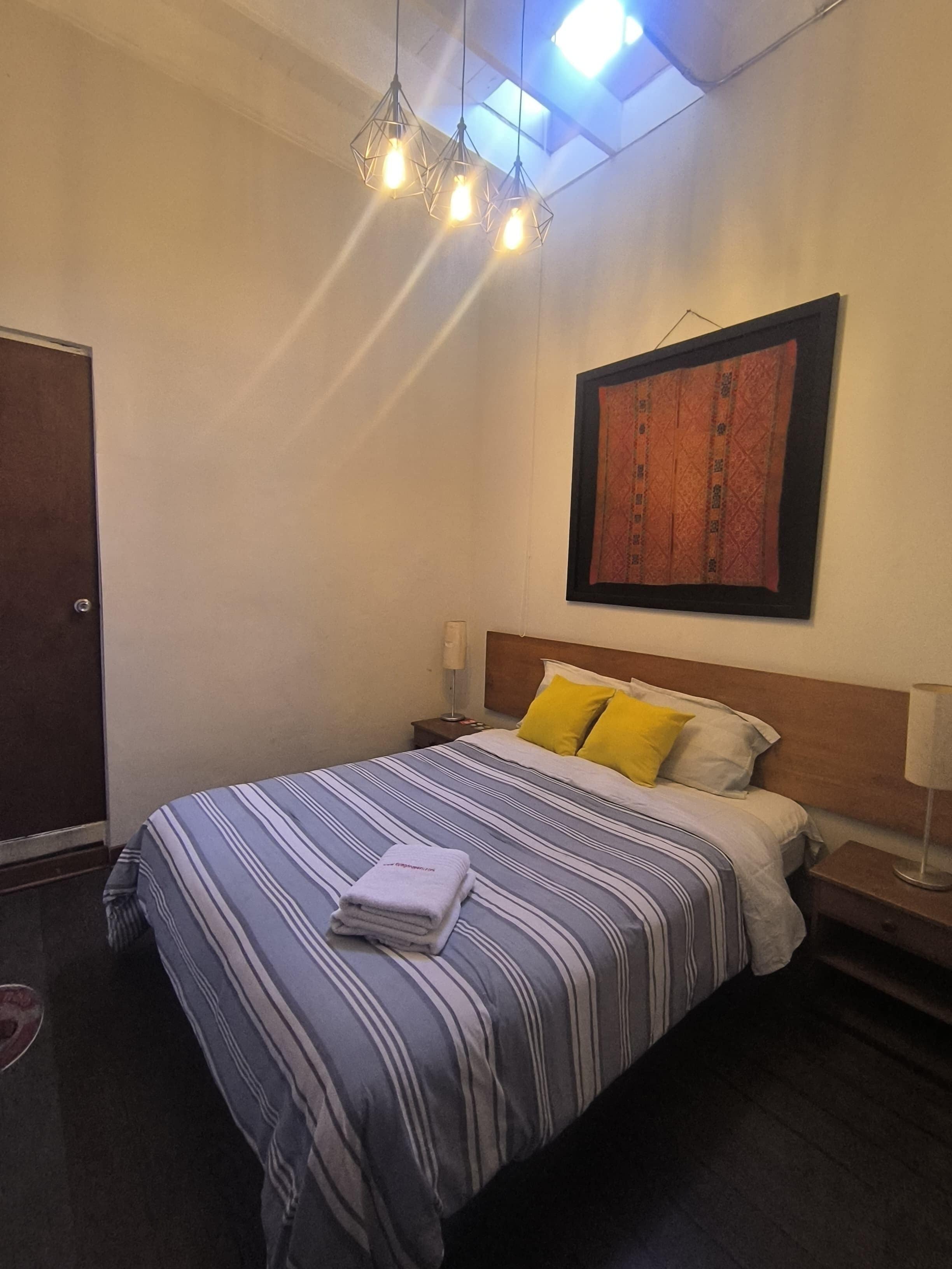 A neatly made bed with striped bedding and two pillows, a bedside table with a lamp, a framed artwork on the wall, and a door to the left.