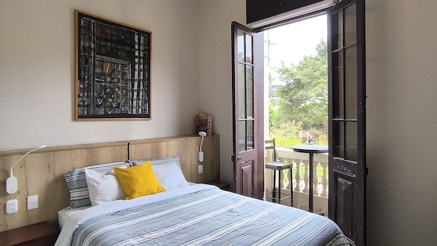 A bedroom with a bed, a wooden headboard, a painting, a bedside table, a nightstand, a wall socket, a power outlet, and a door leading to an outdoor balcony. - Flying Dog Hostel B&B (Lima)