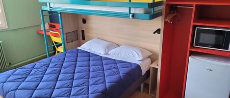 Una cama individual con edredón azul acolchado y almohadas blancas, un refrigerador pequeño blanco y un armario rojo con microondas.