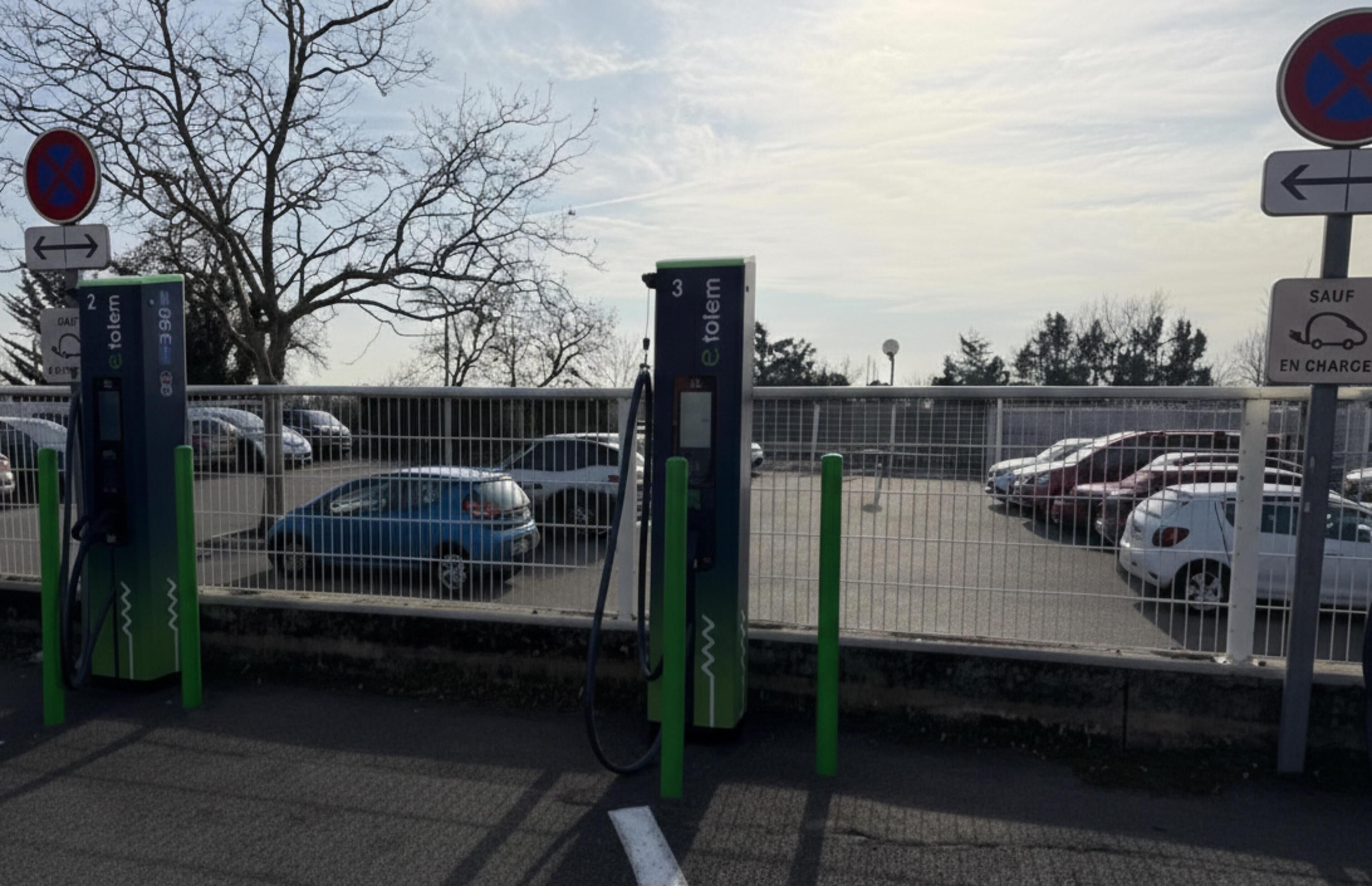 Zona d'aparcament amb estacions de càrrega per vehicles elèctrics, una tanca i diversos cotxes estacionats.