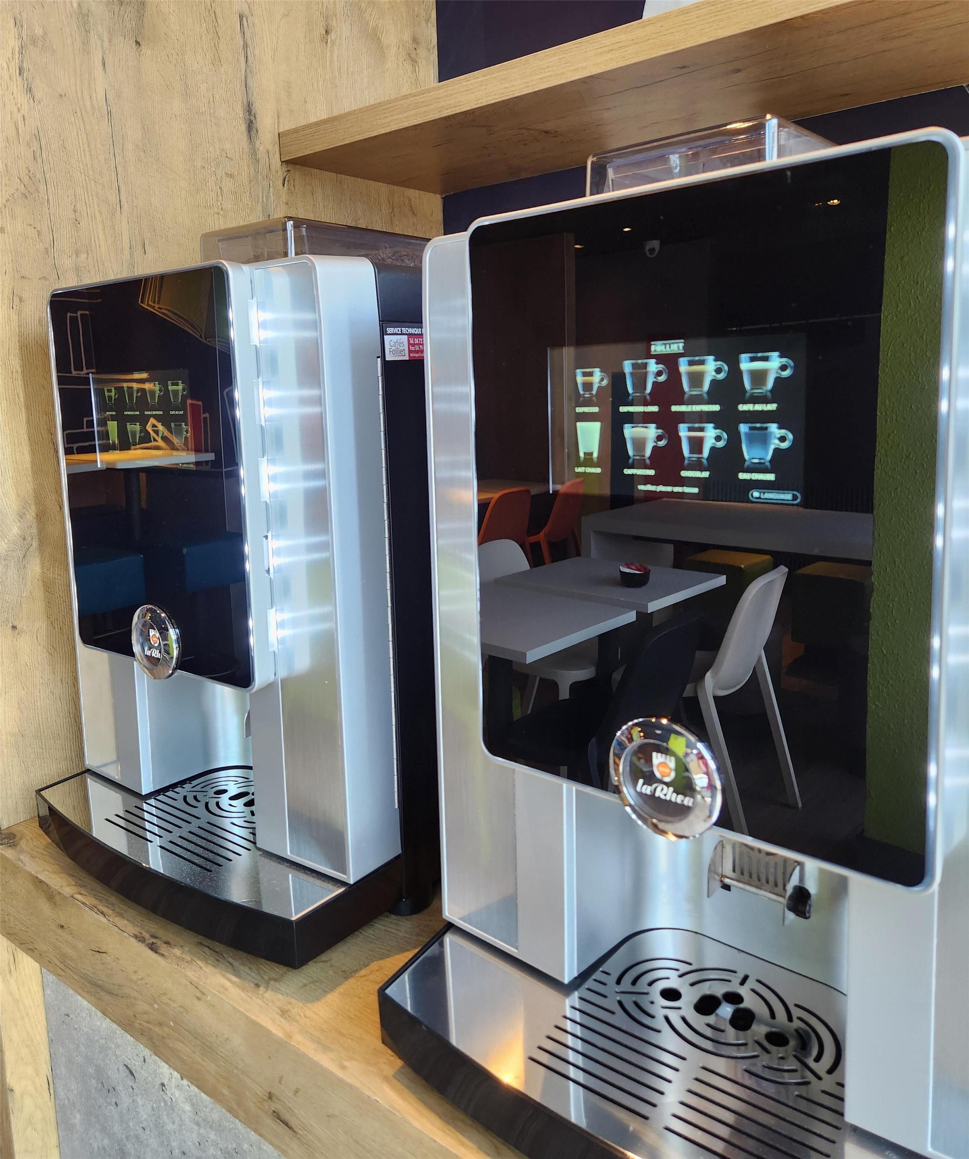 Due macchine da caffè con display digitali del menu, uno scaffale in legno e un'area salotto con tavoli e sedie.
