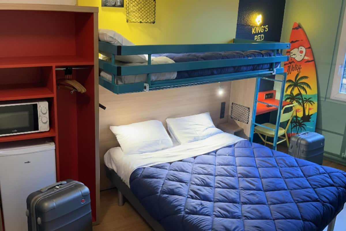 Una cama nido con colcha azul, un refrigerador pequeño, un microondas y una maleta en una habitación con paredes amarillas y una decoración de tabla de surf.