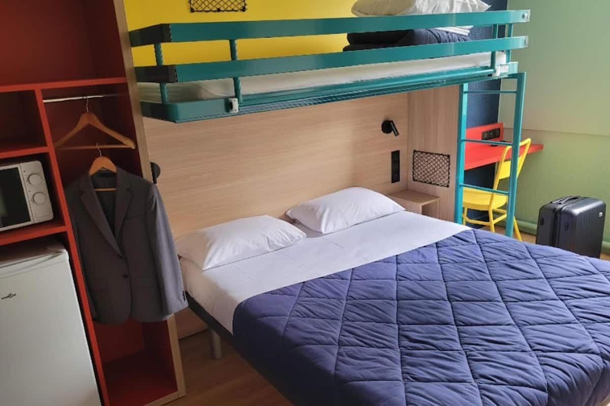 Una cama litera con colcha azul, una mesita de noche pequeña, una silla y un armario con abrigo colgado.