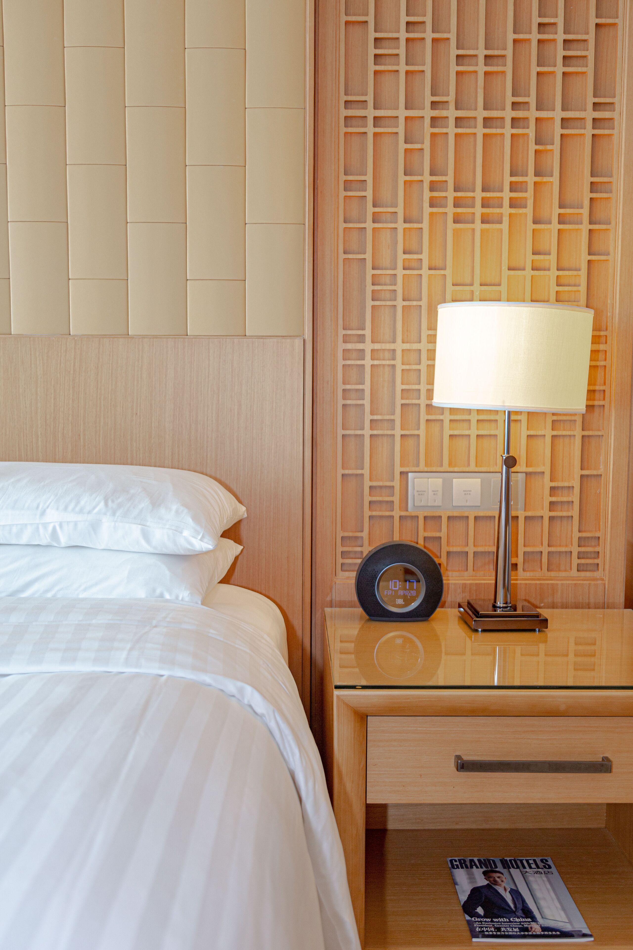A hotel room with a neatly made bed, a bedside table with a lamp, a magazine, and a wooden headboard.