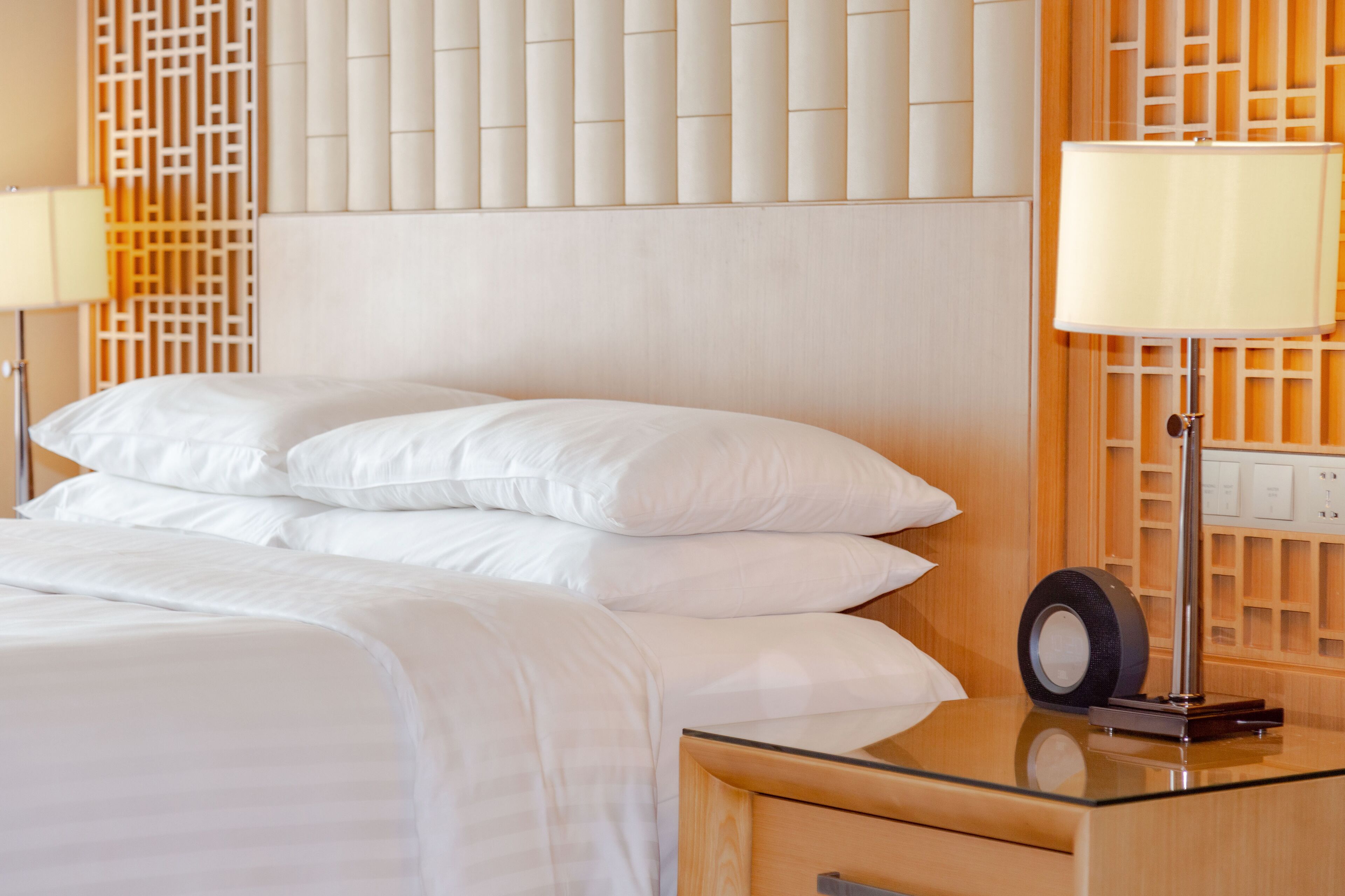A neatly made bed with white linens, a glass side table with a lamp, and a wooden headboard with a decorative panel.