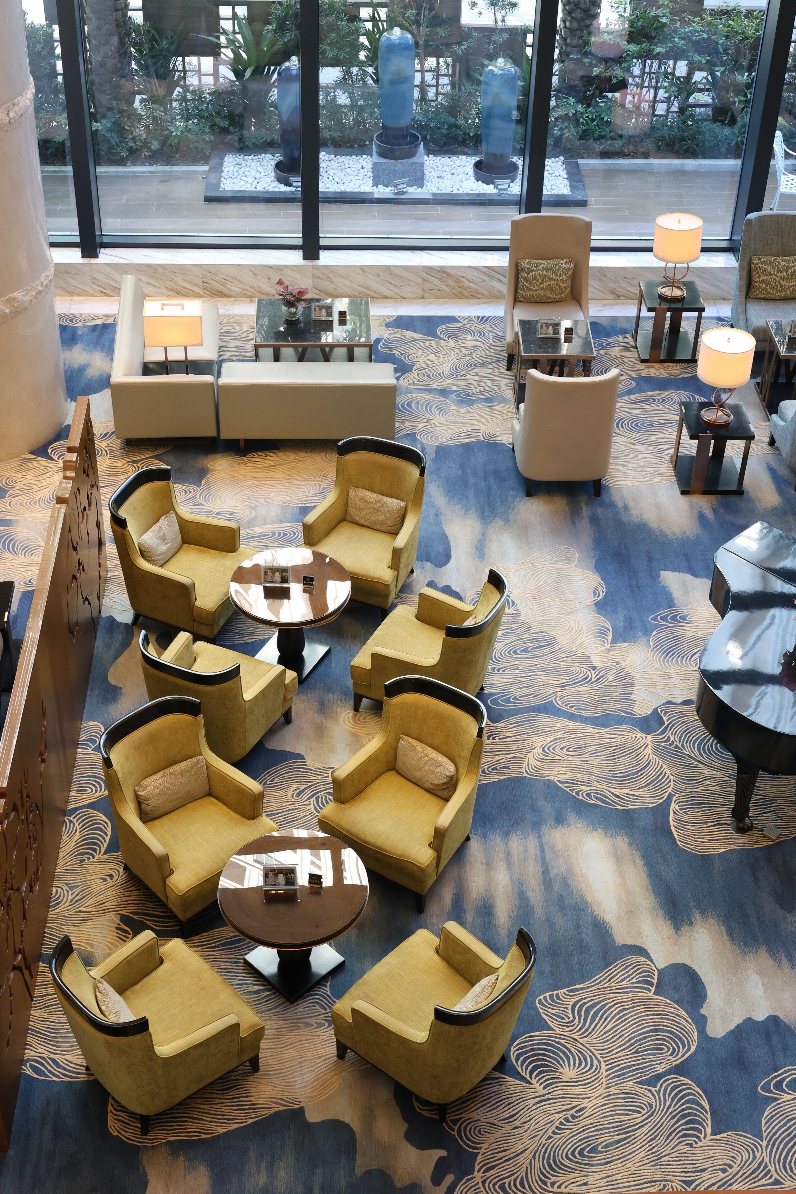 A lobby with a patterned carpet, multiple seating areas including sofas and armchairs, and a view of the outdoor garden through large windows.
