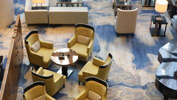 A lobby with a patterned carpet, multiple seating areas including sofas and armchairs, and a view of the outdoor garden through large windows.