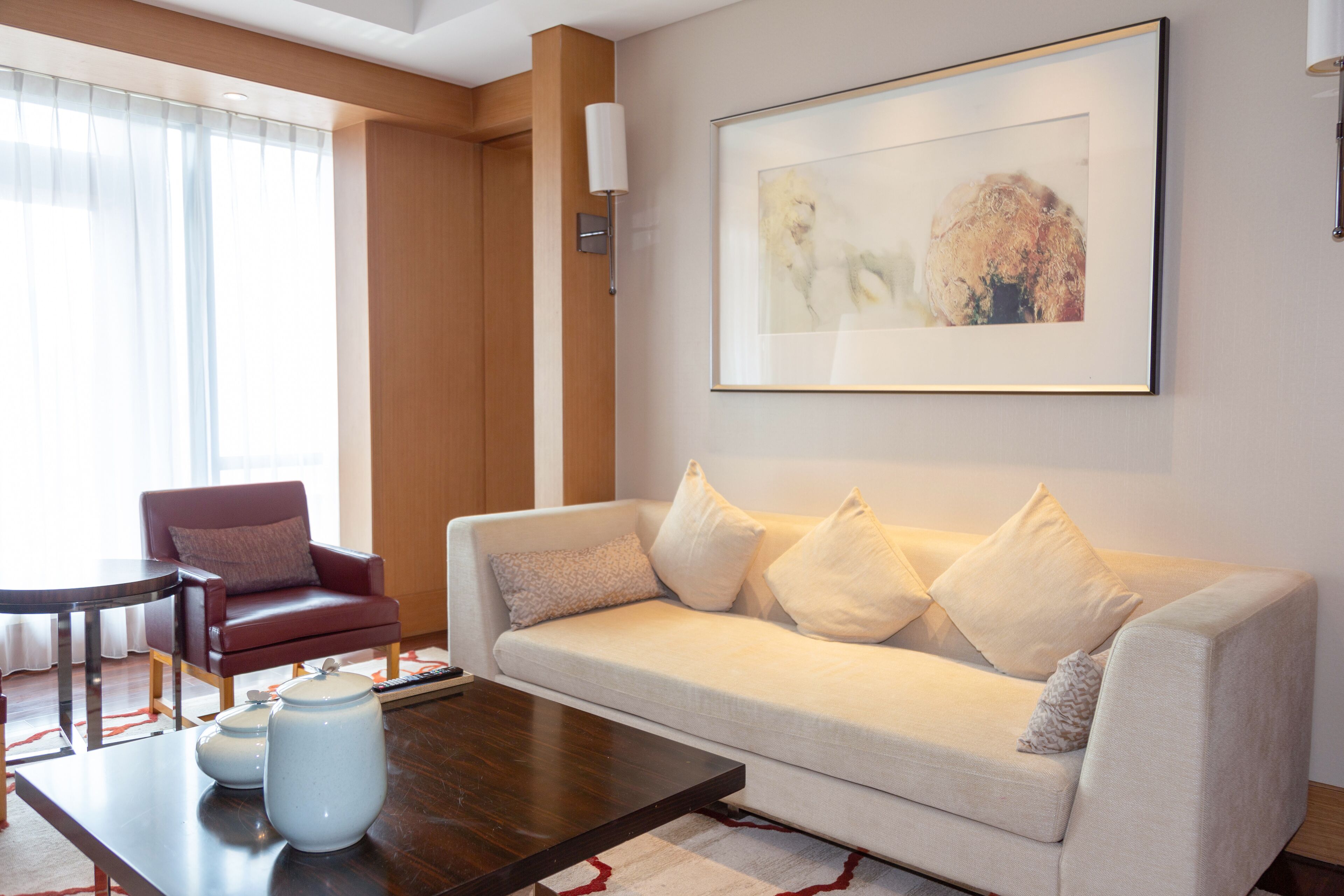 Un salon moderne avec un canapé beige, un fauteuil rouge, une table basse et une œuvre d’art encadrée accrochée au mur.