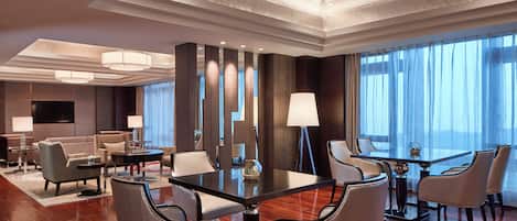 A hotel room with a large dining table, multiple chairs, a sofa, a TV, and a large window with curtains.