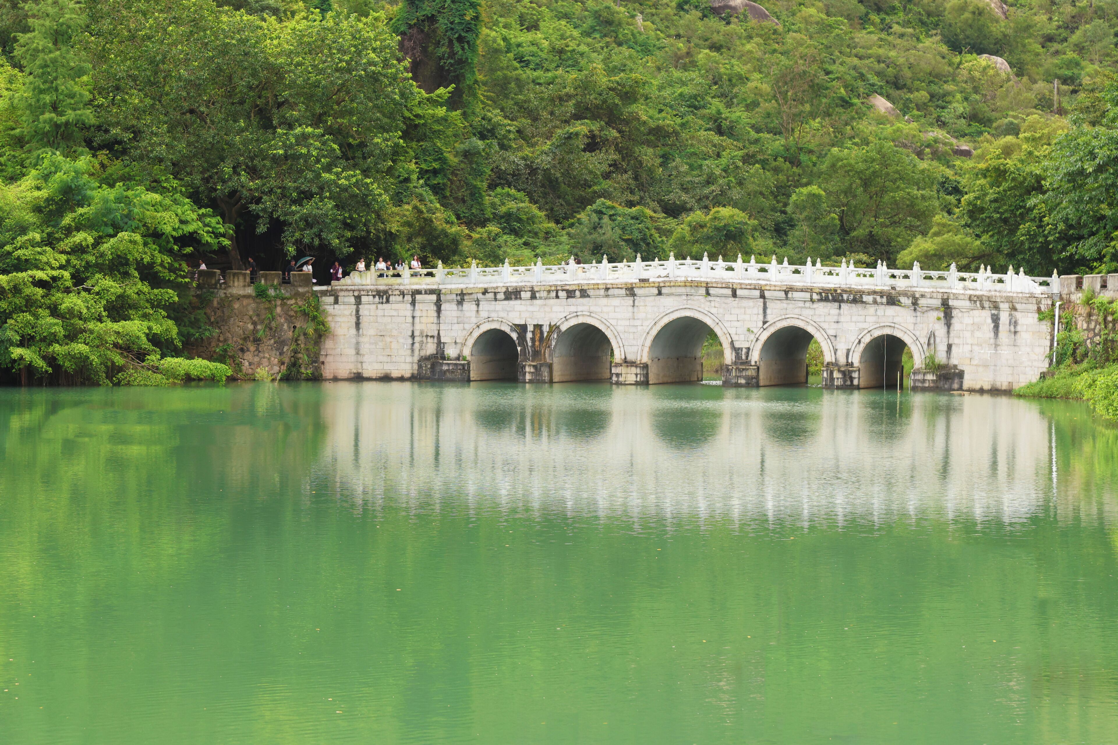 A stone bridge with multiple arches over a calm body of water, surrounded by lush greenery.