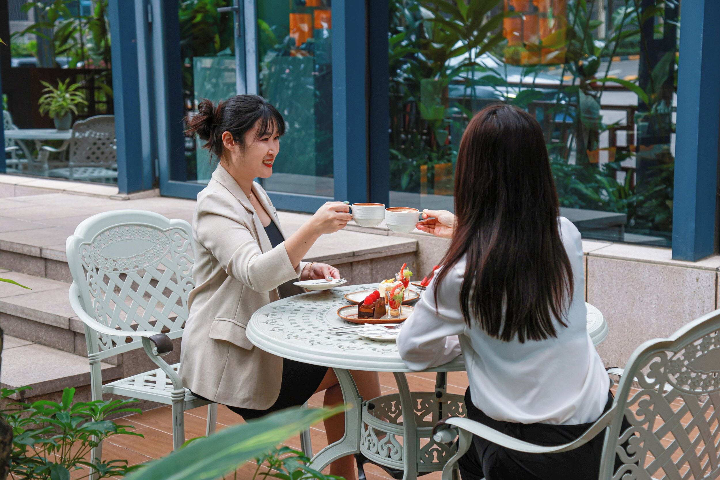 Two people seated at an outdoor table with a glass partition, holding cups, and a plate with food.