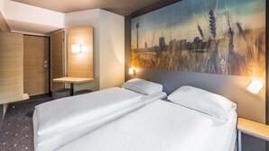 Une chambre d’hôtel avec un lit, un bureau, une chaise, une armoire et un grand tableau représentant un paysage urbain accroché au mur.