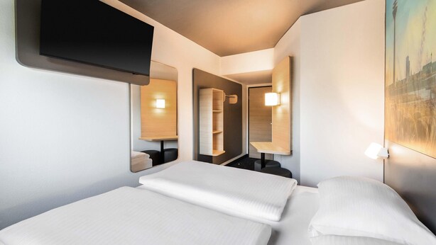 Ein Hotelzimmer mit zwei Betten, einem an der Wand befestigten Flachbildfernseher und einem Stadtbild.