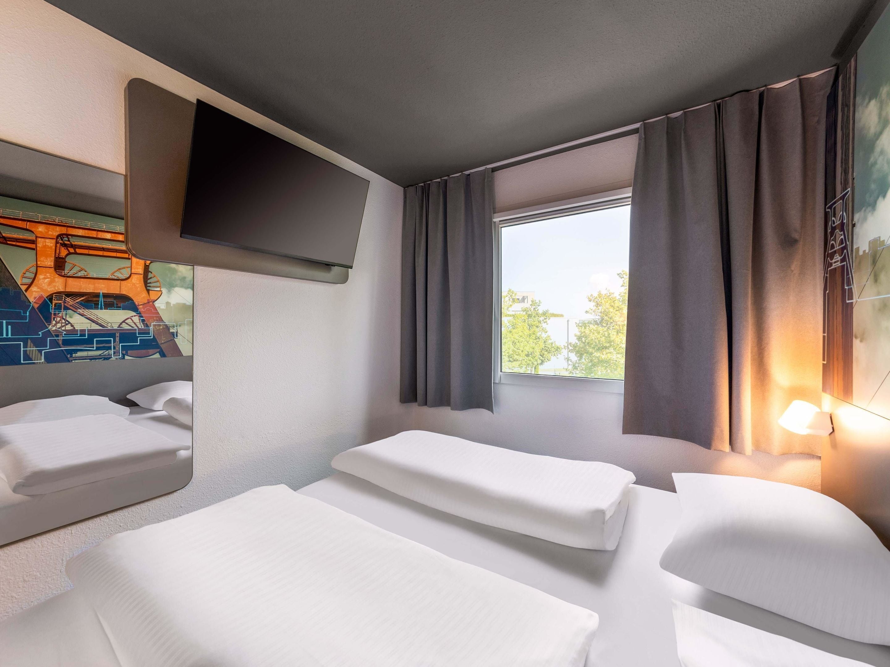A hotel room with two beds, a flat-screen TV mounted on the wall, and a window with curtains.