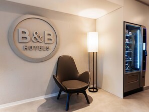Ein Empfangsbereich eines B&B Hotels mit einem modernen Sessel, einem an der Wand befestigten Schild und einem Automaten.