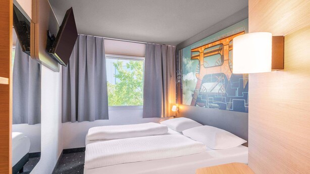 A hotel room with a bed, a TV mounted on the wall, a window with curtains, and a mural of a bridge. -  B&B HOTEL Essen-Nord (Essen)