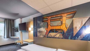 A hotel room with a bed, a desk, a chair, a TV, and a large mural on the wall. -  B&B HOTEL Essen-Nord (Essen)