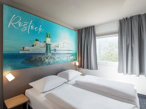 Une chambre d’hôtel comprenant un lit, une table de chevet, une lampe fixée au mur, une fenêtre avec des rideaux et un tableau représentant un phare.