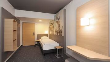 Habitación de hotel moderna con cama, una mesita de noche, una repisa, una lámpara de pared, una estantería de madera y una puerta que da a otra habitación.