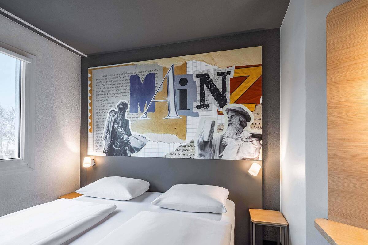 Ein Hotelzimmer mit Bett, einem kleinen Holfnachtkästchen und einem an der Wand hängenden Kunstwerk.