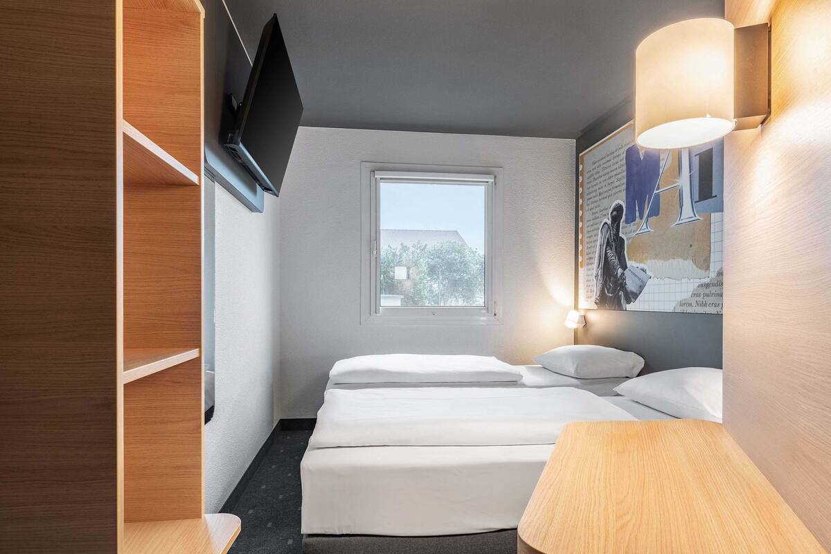 Ein Hotelzimmer mit Bett, Nachttischen, einem an der Wand befestigten Fernseher und einem Fenster mit Blick nach draußen.